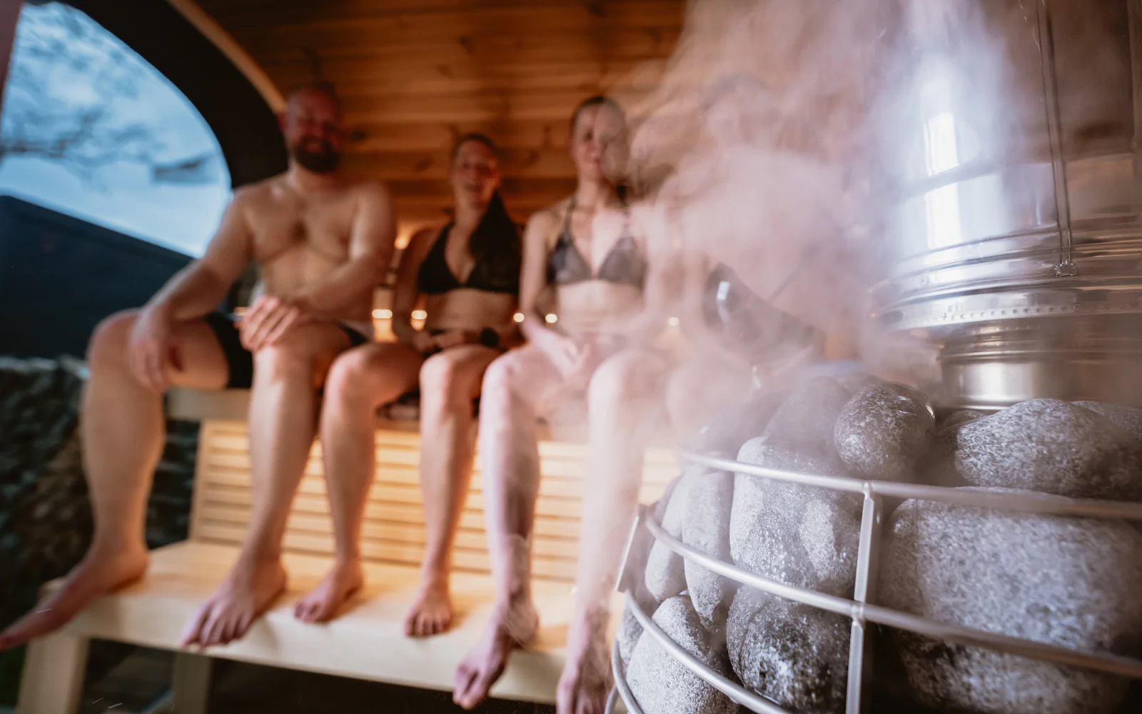 Three people sitting together in a steamy timber sauna beside the stove