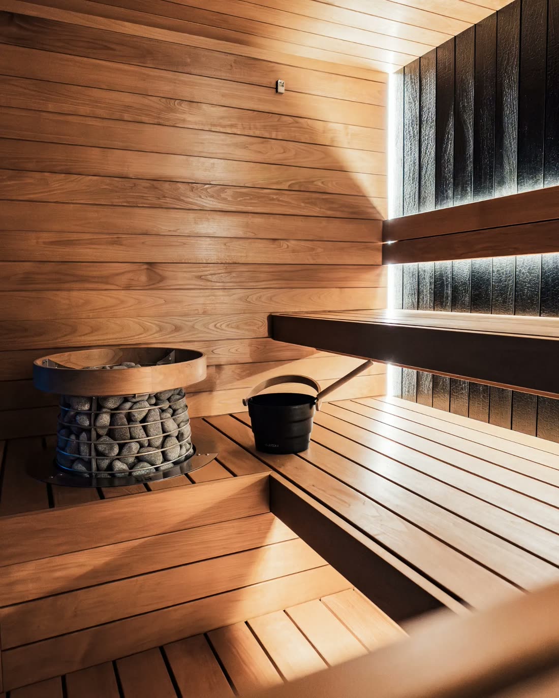 Inside a timber sauna: a basket of hot stones, a black water bucket and slatted benches